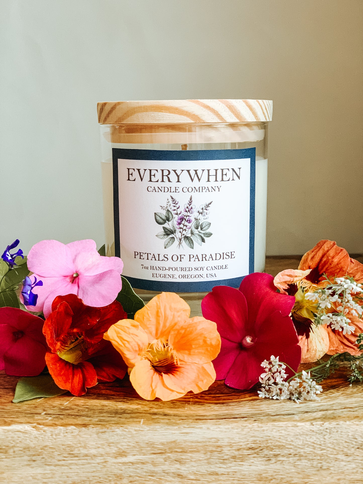 A container candle in a glass jar with pine wood lid, surrounded by fresh summer flowers all brightly colored.